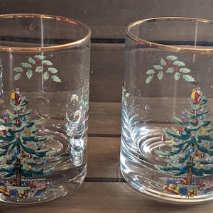 Op de afbeelding: Twee heldere drinkglazen met gouden randen. Elk glas heeft een kerstboomontwerp met ornamenten en een kerstmanfiguur bovenaan. Hulst- en bessenranken zijn ook op het glas geschilderd. De glazen staan op een houten oppervlak.