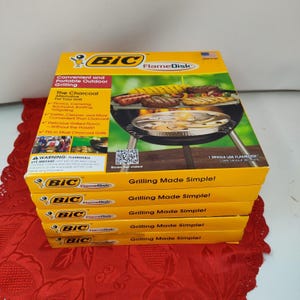 Puede incluir: Una pila de cajas BIC FlameDisk amarillas y negras. Las cajas presentan imágenes de comida a la parrilla y el texto "Grilling Made Simple!" y "Convenient and Portable Outdoor Grilling."