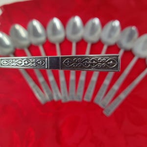 Könnte beinhalten: Ein Set silberner Löffel, angeordnet auf einer roten Oberfläche. Die Löffel haben ein klassisches Design mit ovalen Schalen. Ein Servierutensil mit einem verzierten, gravierten Griff befindet sich im Vordergrund. Der Griff weist ein detailliertes, symmetrisches Muster auf.