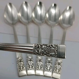 May include: A set of five silver-toned metal spoons featuring a horizontal handle design with a detailed floral relief pattern. The spoons have a smooth, polished bowl and a rectangular end, with one spoon positioned horizontally across the others.