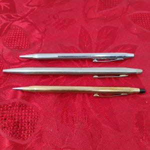 Könnte beinhalten: Drei Stifte vor rotem Hintergrund. Die oberen und mittleren Stifte sind silberfarben, während der untere Stift goldfarben ist. Jeder Stift hat einen Clip und eine Spitze. Die Stifte haben ein schlankes, modernes Design.