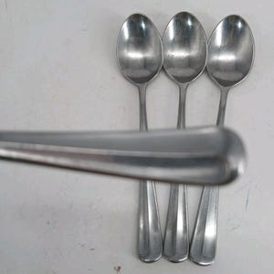 Puede incluir: Tres cucharas de metal de color plateado dispuestas verticalmente con los cuencos hacia arriba. Un cuarto mango de cuchara se coloca horizontalmente en primer plano, ligeramente desenfocado, mostrando un extremo redondeado y una nervadura central.