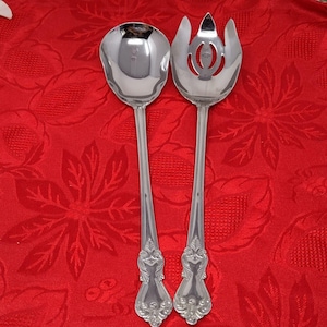 以下が含まれることがあります： 銀色のサービングスプーンとフォークのセット。スプーンは丸みを帯びたボウルで、フォークはスロットデザインです。どちらの器具も、装飾的なディテールが施された長く細いハンドルが付いています。セットは赤い花のテーブルクロスの上に表示されています。