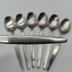 Puede incluir: Un conjunto de siete cucharas de plata. Seis cucharas están en el fondo, con una séptima en primer plano. Las cucharas tienen un diseño sencillo y elegante con cuencos redondeados y mangos largos y delgados. Las cucharas están dispuestas en fila.