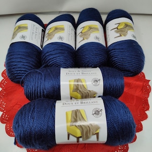 Puede incluir: Un conjunto de madejas de hilo azul marino. Cada madeja lleva la etiqueta "Soft & Shiny Doux et Brillant". El hilo está dispuesto sobre un tapete rojo y calado. La imagen muestra la textura y el color del hilo.