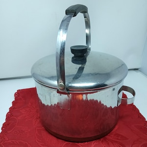 Peut inclure: Une marmite cylindrique en acier inoxydable avec une finition polie, dotée d'un couvercle assorti avec un bouton noir. La marmite comprend une grande poignée métallique arquée avec une prise noire et une petite poignée latérale circulaire.