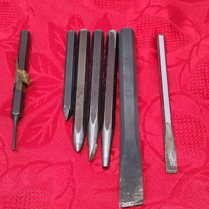 Peut inclure: Ensemble d'outils métalliques, comprenant des poinçons et des burins, disposés sur un tissu rouge. Les outils varient en taille et en forme, certains ayant des pointes et d'autres des bords biseautés plats. Les outils sont fabriqués aux États-Unis.