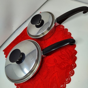 Puede incluir: Dos cacerolas de acero inoxidable con asas y tapas negras. Las cacerolas están sobre un mantel individual rojo estampado. Las tapas tienen pomos redondos negros. Una cacerola tiene un asa larga y la otra, corta.