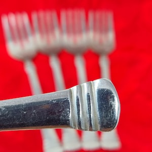 May include: Close-up of a silver fork handle with a ridged design, in focus against a blurred background of additional forks. The forks are set against a vibrant red backdrop, creating a striking contrast.