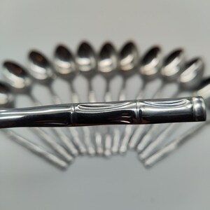 Könnte beinhalten: Eine Ansammlung glänzender, silberner Löffel, die fächerförmig angeordnet sind. Die Löffel haben ein schlichtes, elegantes Design mit einer glatten, reflektierenden Oberfläche. Die Griffe haben eine bambusartige Textur, die dem Besteck einen Hauch von Natürlichkeit verleiht.