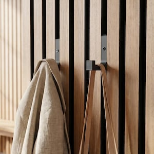 Könnte beinhalten: Ein beigefarbener Leinenmantel und eine Canvas-Tragetasche hängen an schwarzen Metallhaken an einer Holzlattenwand. Die Wand hat vertikale Holzlatten in abwechselnden hellen und dunklen Farbtönen. Im Hintergrund ist eine Holzbank zu sehen.