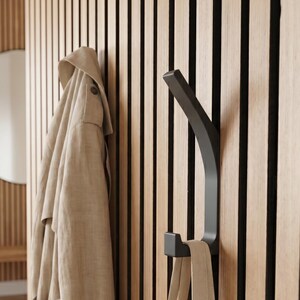 Può includere: Un gancio da parete in metallo nero opaco dal design curvo montato su un pannello a listelli di legno verticali. Un trench beige è appeso al gancio superiore, mentre una borsa tote in tela beige è appesa al gancio inferiore della struttura.