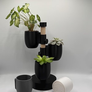 Könnte beinhalten: Ein schwarzer Pflanzenständer mit drei schwarzen Töpfen und einem Holzstift. Der Ständer hat eine runde Basis und einen weißen Topf mit Abzugslöchern. Eine grüne Pflanze befindet sich im oberen Topf, eine Sukkulente im mittleren Topf und eine Blattpflanze im unteren Topf.