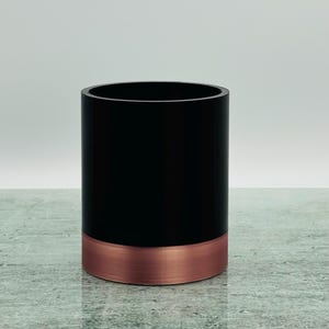 May include: A black cylindrical container with a copper-coloured base. The container is on a textured surface, with a plain, light grey background. The design is simple and modern.