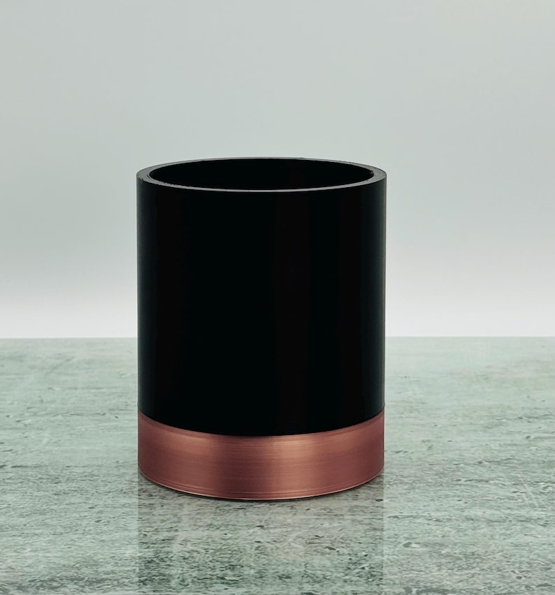 May include: A black cylindrical container with a copper-coloured base. The container is on a textured surface, with a plain, light grey background. The design is simple and modern.