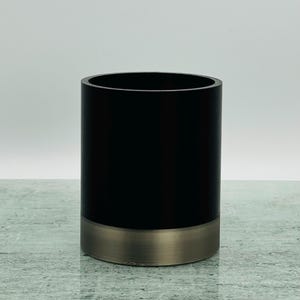 May include: A cylindrical container with a matte black upper section and a brushed metal base. The container has a smooth, polished finish and a simple, modern design. The interior is not visible.