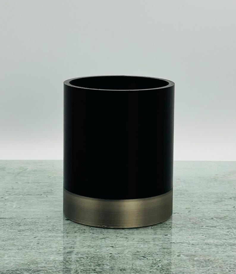 May include: A cylindrical container with a matte black upper section and a brushed metal base. The container has a smooth, polished finish and a simple, modern design. The interior is not visible.