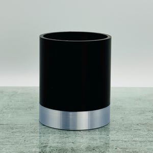 May include: A cylindrical container. The upper section is black, and the base is silver. The container has a smooth, matte finish. The silver base provides a metallic accent. The height is approximately 10 cm.