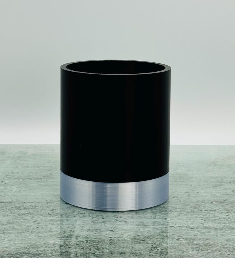 May include: A cylindrical container. The upper section is black, and the base is silver. The container has a smooth, matte finish. The silver base provides a metallic accent. The height is approximately 10 cm.