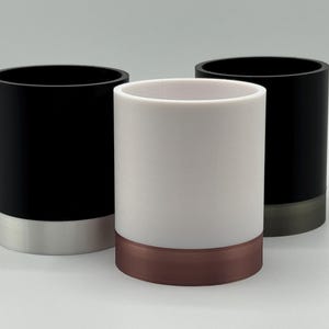 May include: Three cylindrical containers. Two are black with silver-coloured bases, and one is white with a copper-coloured base. The containers are of equal height and have a smooth, matte finish. The background is a solid, light grey.