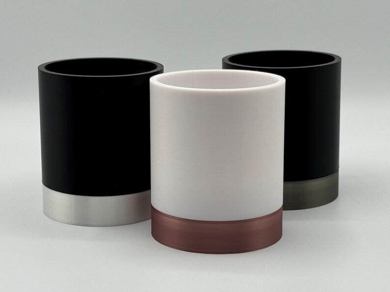 May include: Three cylindrical containers. Two are black with silver-coloured bases, and one is white with a copper-coloured base. The containers are of equal height and have a smooth, matte finish. The background is a solid, light grey.
