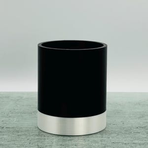 May include: A cylindrical container. The upper section is black, and the base is silver. The container has a smooth finish and a simple, modern design. It is suitable for holding various items.