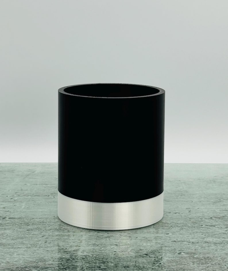 May include: A cylindrical container. The upper section is black, and the base is silver. The container has a smooth finish and a simple, modern design. It is suitable for holding various items.