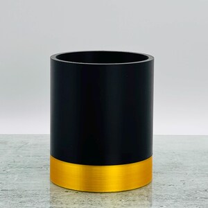 May include: A cylindrical container, black above and gold-coloured at the base, with a brushed metal effect. The container is empty and stands on a reflective surface. The design is minimalist and modern.