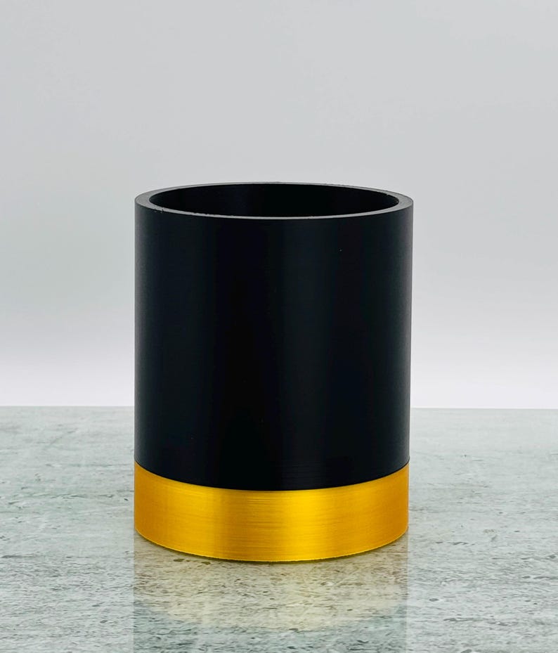May include: A cylindrical container, black above and gold-coloured at the base, with a brushed metal effect. The container is empty and stands on a reflective surface. The design is minimalist and modern.
