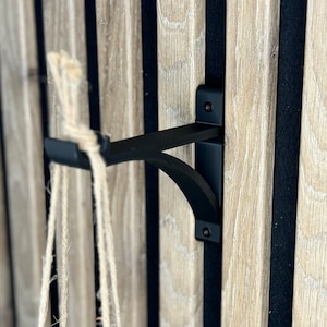 Op de afbeelding: Zwarte metalen plantenhangerbeugel gemonteerd op een houten muur met zwarte verticale strepen. De beugel heeft een gebogen arm en een haak aan het einde. Een touw van natuurlijke vezels hangt aan de haak.
