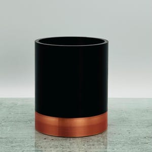 May include: A black cylindrical container with a copper-coloured base. The container is on a grey surface. The design is simple and modern, suitable for various uses.