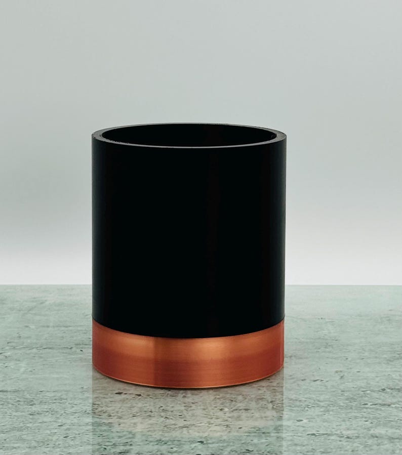 May include: A black cylindrical container with a copper-coloured base. The container is on a grey surface. The design is simple and modern, suitable for various uses.