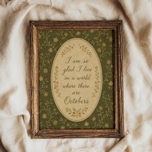 May include: A rectangular dark brown wood-textured frame containing a print with a dark green background featuring a delicate floral pattern. In the center, an oval cream-colored field displays the text: I am so glad I live in a world where there are Octobers.