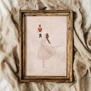 Könnte beinhalten: Eine gerahmte Illustration, die eine Ballerina in einem hellrosa Tutu zeigt, die nach einem kleinen, rot-schwarzen Nussknacker-Soldaten greift. Das Kunstwerk befindet sich in einem rechteckigen, dunkelbraunen Holzrahmen mit sichtbarer Maserung.