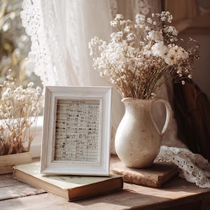 Puede incluir: Un marco de fotos de madera blanca que contiene una tarjeta de biblioteca antigua con el texto DATE DUE, junto a una jarra de cer&aacute;mica color crema llena de flores blancas secas. El marco descansa sobre un libro viejo, creando un bodeg&oacute;n.