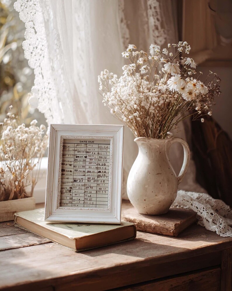 Puede incluir: Un marco de fotos de madera blanca que contiene una tarjeta de biblioteca antigua con el texto DATE DUE, junto a una jarra de cer&aacute;mica color crema llena de flores blancas secas. El marco descansa sobre un libro viejo, creando un bodeg&oacute;n.