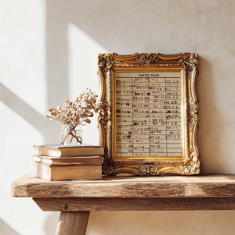 Puede incluir: Una tarjeta de biblioteca antigua con fechas selladas, enmarcada en un marco dorado ornamentado. La tarjeta muestra columnas de fechas y el texto DATE DUE en la parte superior. Junto al marco hay un peque&ntilde;o jarr&oacute;n de cristal con flores secas.