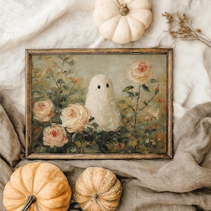 May include: A framed painting of a white ghost among pink roses. The artwork has a vintage aesthetic, with a wooden frame. Two pumpkins and a small gourd are arranged around the painting, suggesting a fall or Halloween theme.
