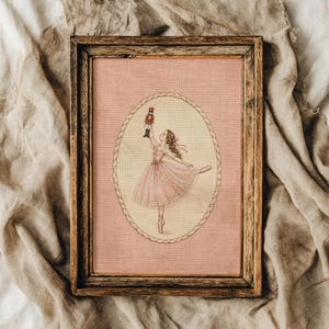 Op de afbeelding: Een ingelijst kruissteek borduurwerk met een ballerina in een roze tutu die een notenkrakerfiguur vasthoudt. Het ontwerp is gecentreerd binnen een ovale, touwachtige rand op een oudroze stoffen achtergrond, in een rustieke houten lijst.