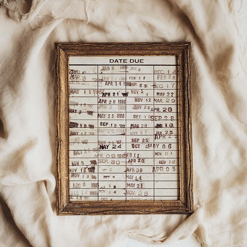 Puede incluir: Un marco de madera rectangular que contiene una tarjeta de biblioteca vintage con el t&iacute;tulo DATE DUE. La tarjeta presenta una cuadr&iacute;cula con numerosas fechas estampadas en tinta negra, incluyendo varios meses y a&ntilde;os de los 80, en columnas.