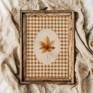 May include: A rectangular wooden frame containing a print with a brown and white gingham pattern background. Centred on the print is an oval shape featuring a single, detailed illustration of a golden-brown maple leaf with visible veins.