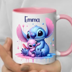 K&ouml;nnte beinhalten: Eine wei&szlig;e Keramiktasse mit rosafarbenem Rand und Henkel. Die Tasse zeigt eine farbenfrohe Illustration von Stitch und Angel aus dem Disney-Film Lilo & Stitch. Der Text "Emma" ist auf die Tasse gedruckt.