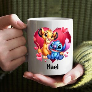 Puede incluir: Taza de cer&aacute;mica blanca con una ilustraci&oacute;n colorida de Simba de El Rey Le&oacute;n y Stitch de Lilo & Stitch dentro de un coraz&oacute;n rojo. El texto "Mael" est&aacute; impreso debajo de la imagen.