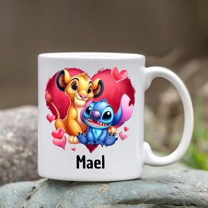 Puede incluir: Taza de cer&aacute;mica blanca con un dise&ntilde;o de coraz&oacute;n rojo que presenta un le&oacute;n de dibujos animados y Stitch de Lilo & Stitch. El coraz&oacute;n tiene acentos rosados y el nombre "Mael" est&aacute; impreso debajo del dise&ntilde;o.