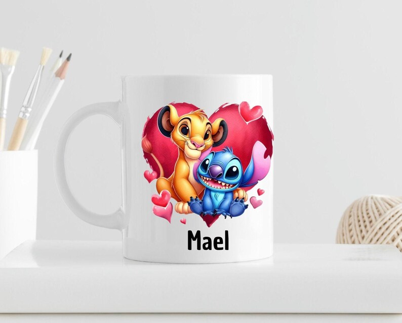 Puede incluir: Taza de cer&aacute;mica blanca con una ilustraci&oacute;n colorida de Simba de El Rey Le&oacute;n y Stitch de Lilo & Stitch. Los personajes est&aacute;n dentro de un coraz&oacute;n rojo con corazones rosas alrededor. La taza tiene el nombre "Mael" impreso en la parte inferior.