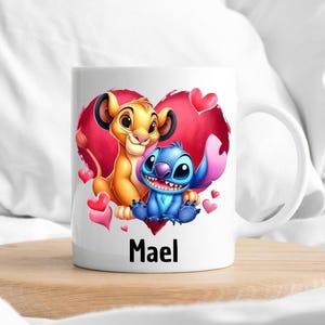 Puede incluir: Taza de cer&aacute;mica blanca con un dise&ntilde;o de coraz&oacute;n rojo que presenta un le&oacute;n de dibujos animados y Stitch de Lilo & Stitch. El coraz&oacute;n tiene corazones rosas y blancos a su alrededor. La taza tiene el nombre "Mael" impreso en la parte inferior.