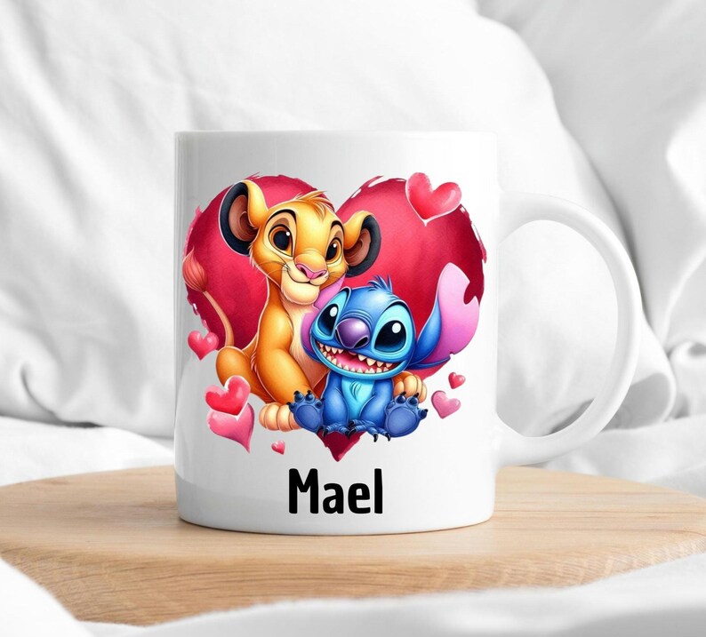 Puede incluir: Taza de cer&aacute;mica blanca con un dise&ntilde;o de coraz&oacute;n rojo que presenta un le&oacute;n de dibujos animados y Stitch de Lilo & Stitch. El coraz&oacute;n tiene corazones rosas y blancos a su alrededor. La taza tiene el nombre "Mael" impreso en la parte inferior.