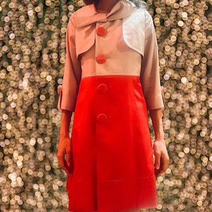 May include: A two-tone coat with a red skirt and a light beige upper section. The upper section has a light beige collar and sleeves, with three red buttons down the front. The coat is knee-length.