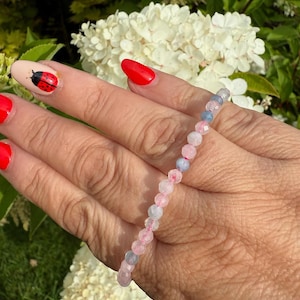 May include: A delicate beaded bracelet featuring alternating pink and blue gemstones. The bracelet is displayed against a background of green foliage and white flowers. The gemstones are small and round, creating a subtle and elegant accessory.