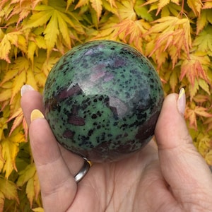 May include: A green and pink sphere with black speckles. The sphere is made of polished stone and has a smooth, shiny surface.